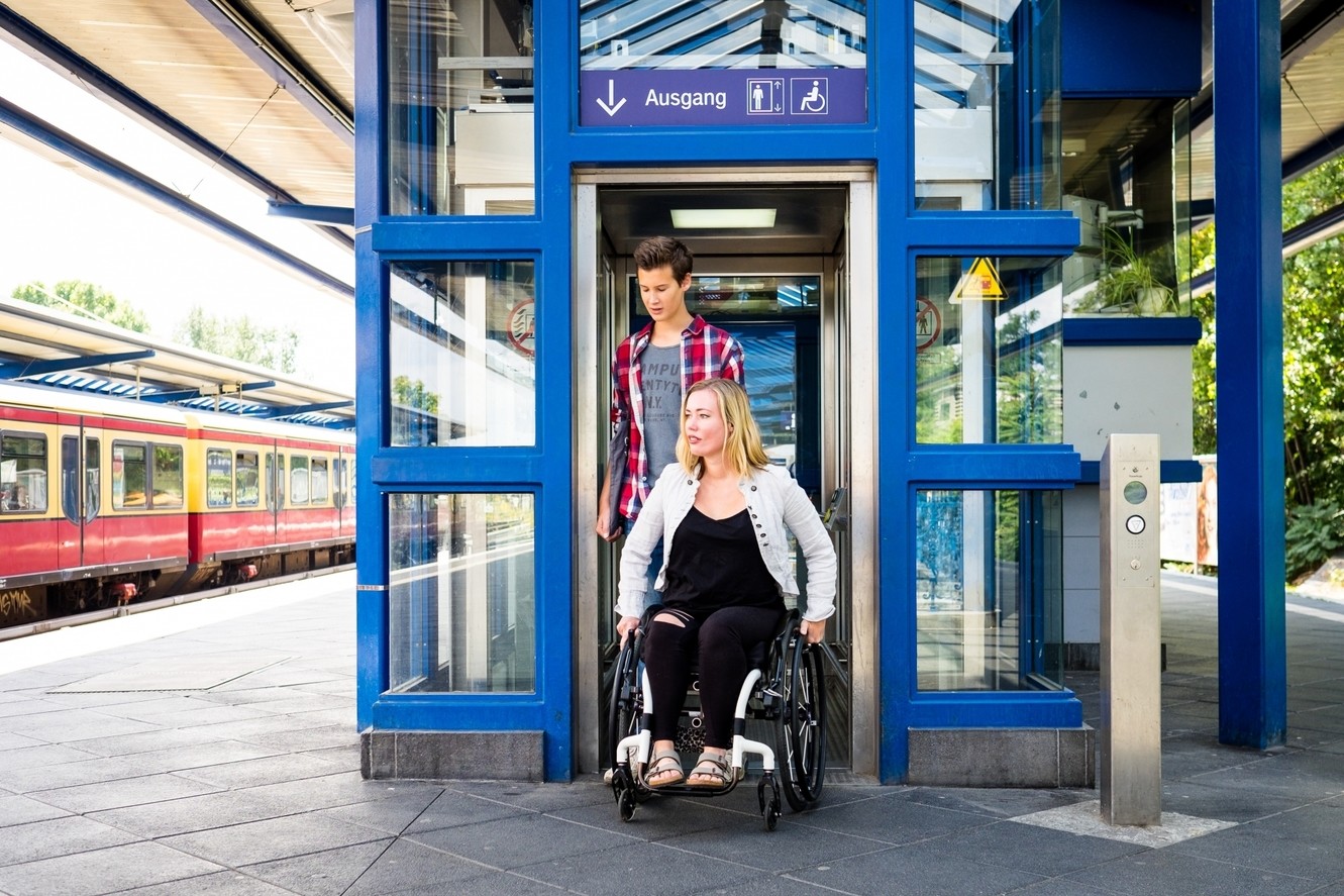 Photo of a woman in a wheelchair, accompanied by a man, leaving an elevator at the suburban railroad.