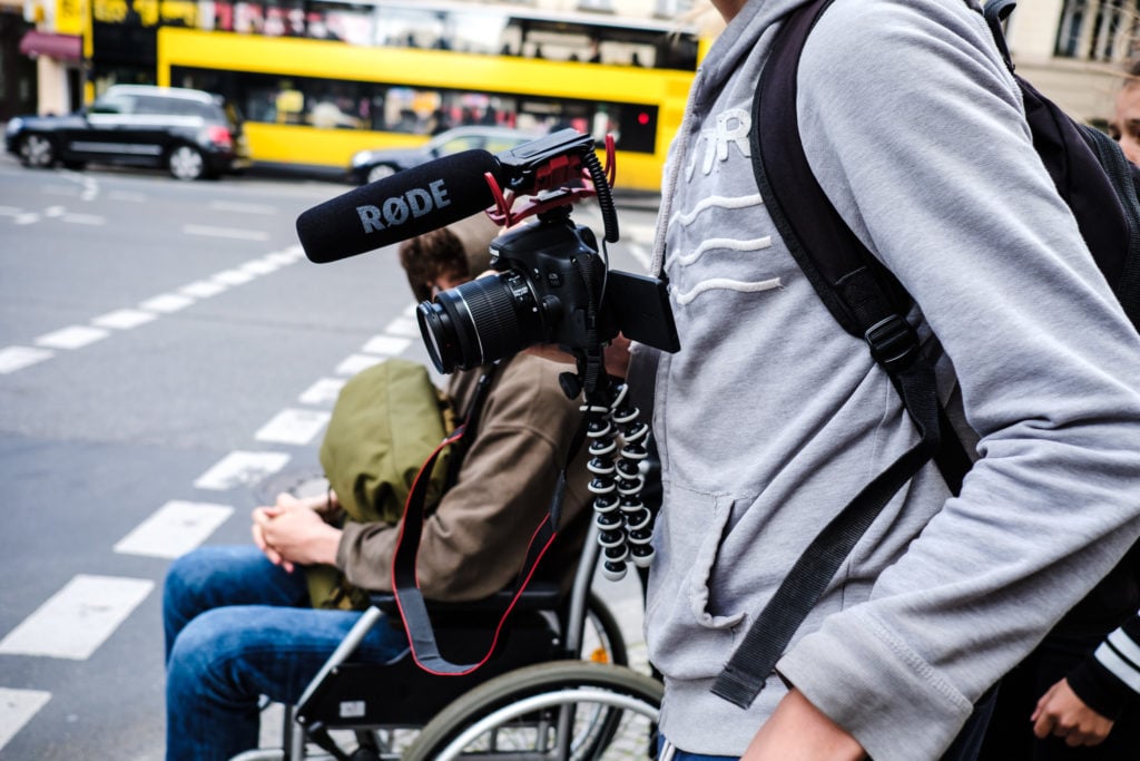 A person with a video camera. In the background a person in a wheelchair