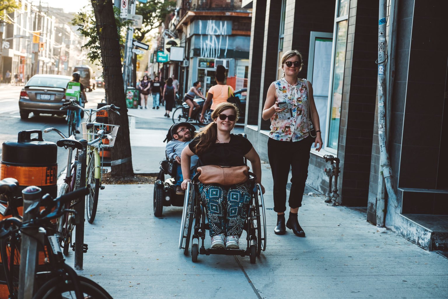 Two wheelchair users and a pedestrian are walking down the street.