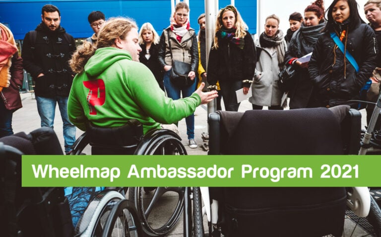 A person sitting in a wheelchair explains something to bystanders. Text: Wheelmap Ambassador Program 2021