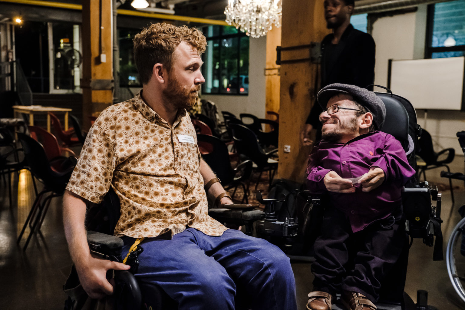 Raul Krauthausen im Rollsuhl mit einem anderen Rollstuhlfahrer unterhalten sich in einem leeren Event-Raum. Im Hintergrund stühle. Raul lächelt.