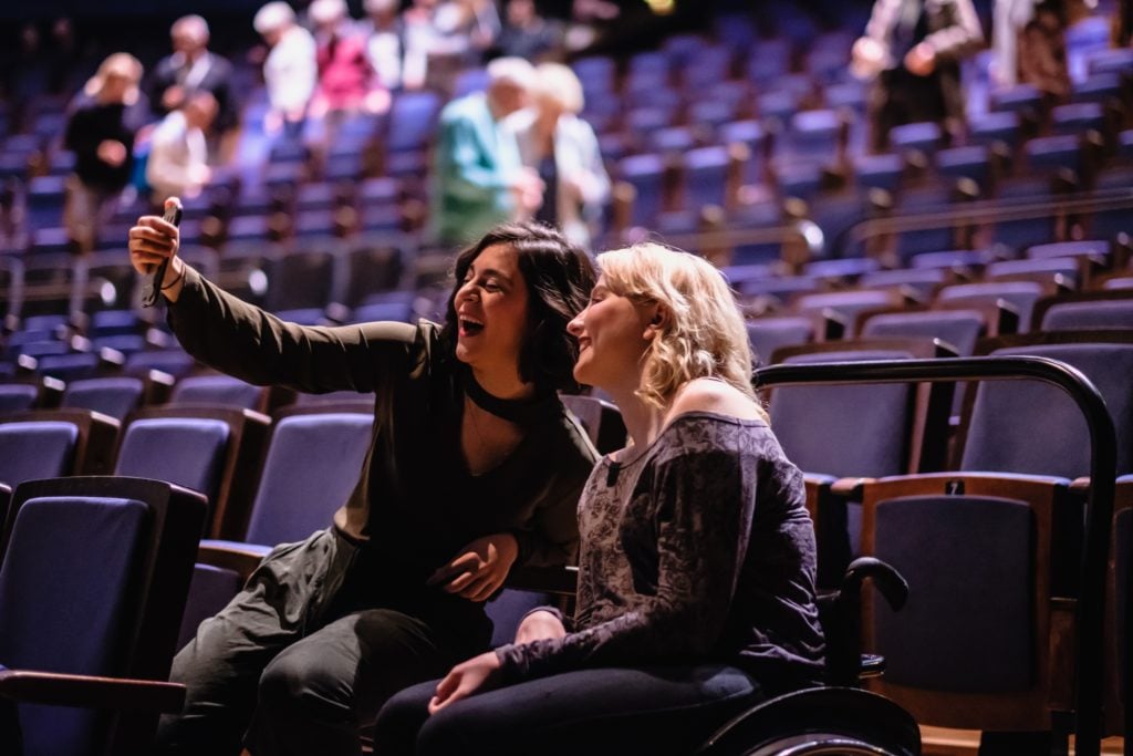 Zwei Frauen, davon eine mit Rollstuhl, sitzen in den Sesseln im Saal des Friedrichstadt Palastes . Sie wirken vergnügt und machen ein gemeinsames Selfie-Foto mit dem Smartphone während im Hintergrund weitere Besucher*innen ihre Plätze einnehmen.