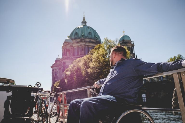 Ein Mann im Rollstuhl sitzt in der Sonne und schaut hinter sich über die Spree auf den Berliner Dom.