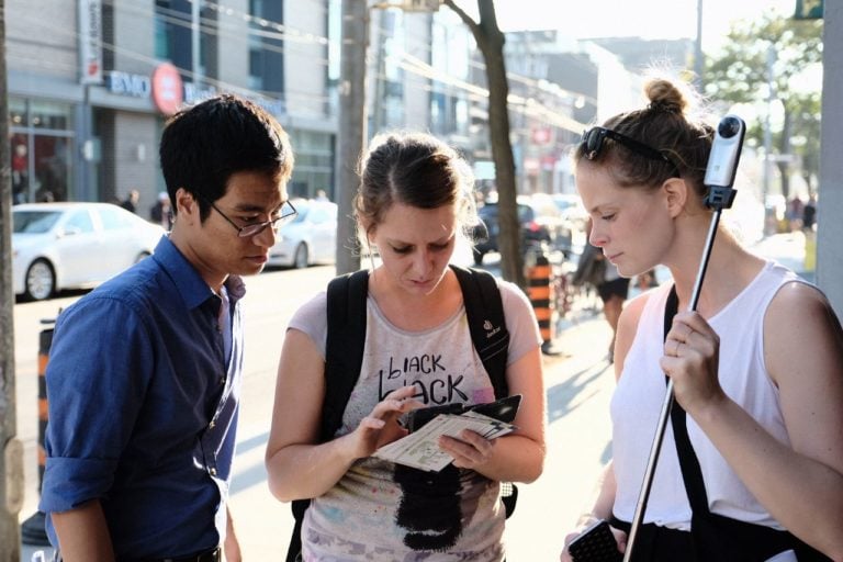 Drei Personen, die an einem Mapping Event teilenehmen. Sie schauen konzentriert gemeinsam auf ein Smartphone. Es ist sehr sommerlich und städtisch.