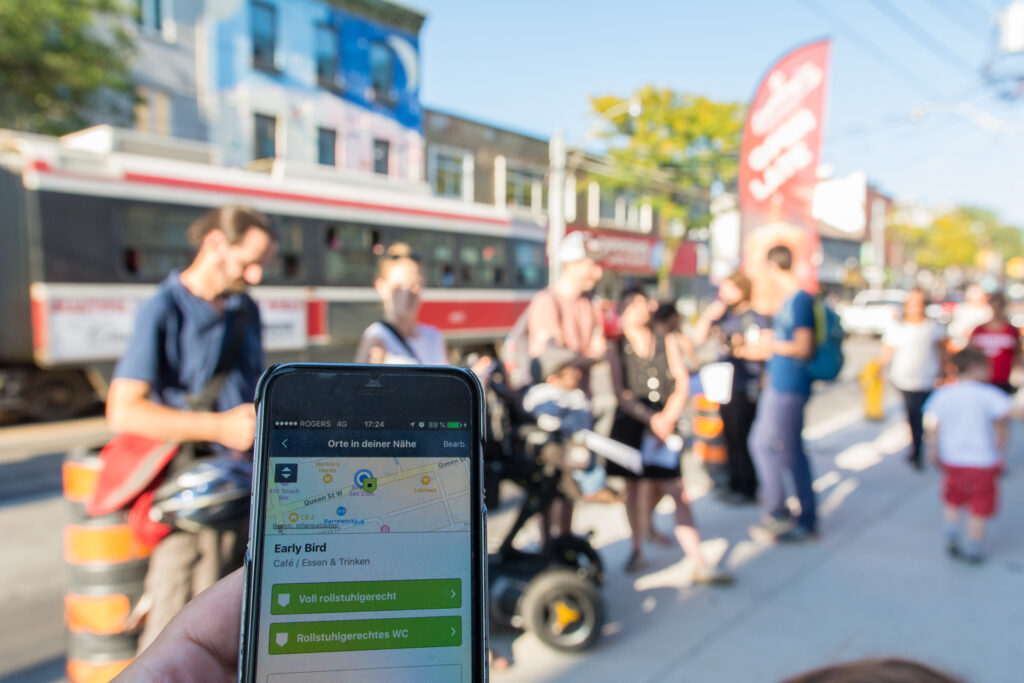 Ein Smartphone mit der Wheelmap-App, im Hintergrund sieht man eine Gruppe von Menschen, die bei Sonnenschein an einer belebten Straße stehen.