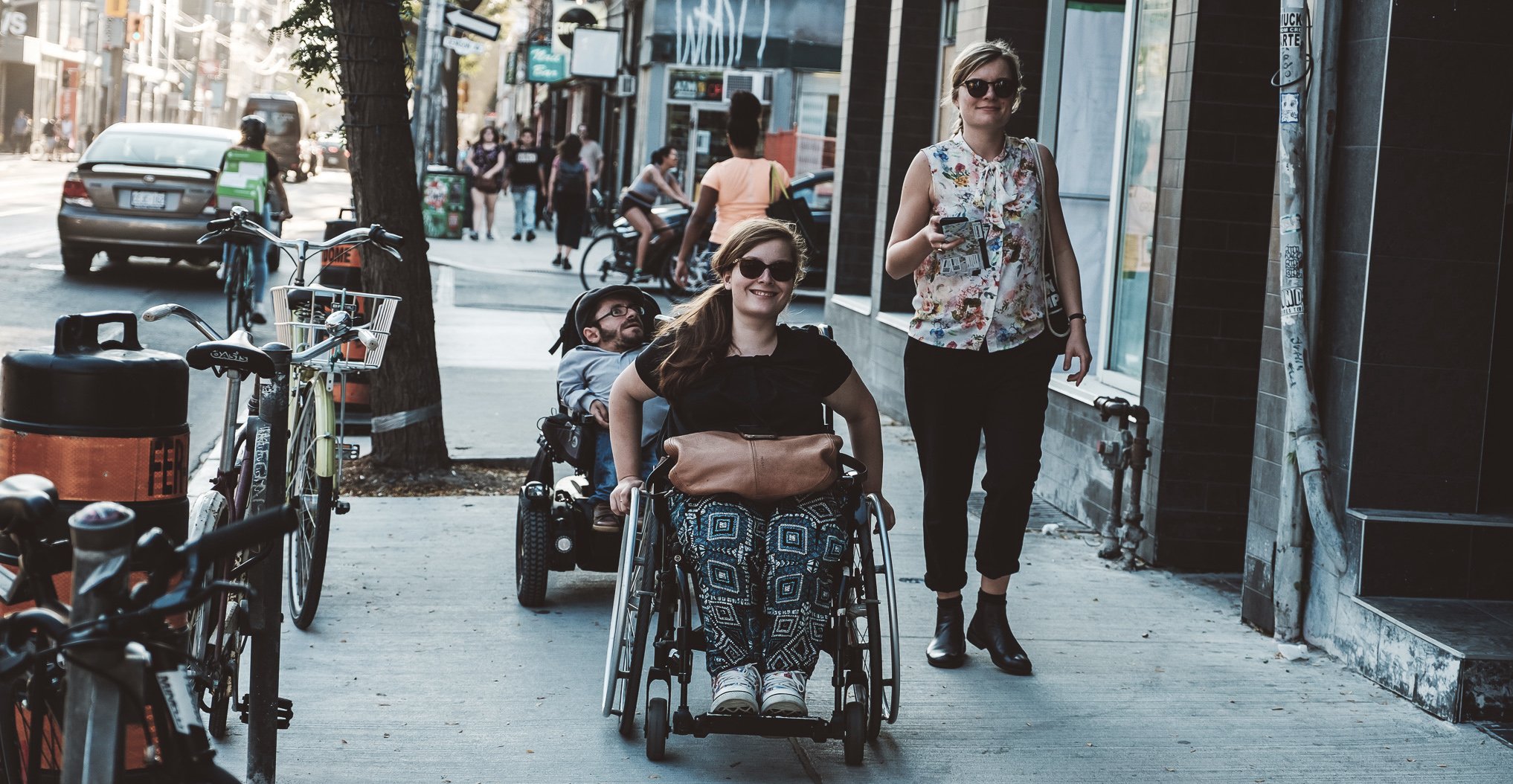 Eine Frau und ein Mann mit Rollstuhl und eine Fußgängerin gehen gemeinsam die Straße entlang
