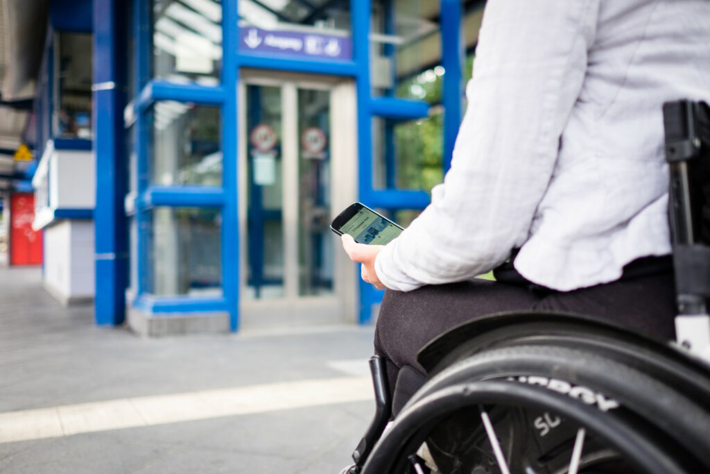 Eine Person im Rollstuhl hat eine Handy in der Hand und steht vor einem Aufzug.